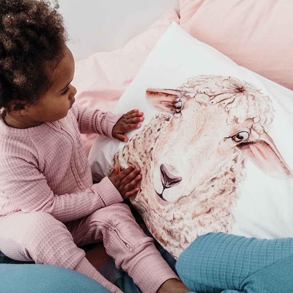 young girl in pink sleepsuit looking at sheep artwork cotton pillowcase on bed